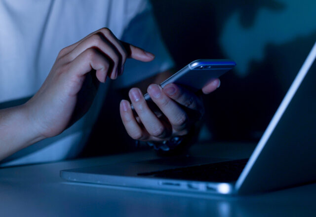 close up young man hand holding smartphone for access by unlock on screen and using laptop for register website on internet in the late night , internet addiction concept