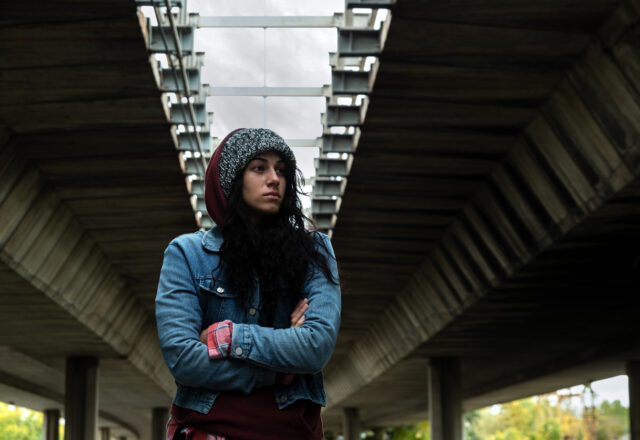 Young depressed homeless girl or woman standing alone under the bridge on the street on the cold weather feeling anxious abandoned and freezing selective focus