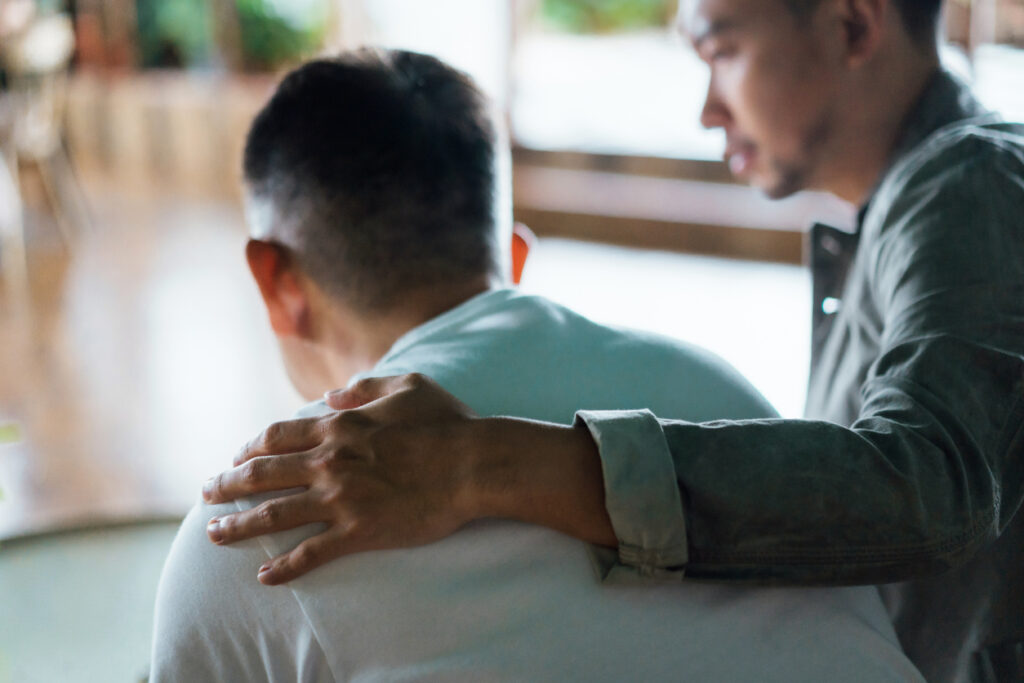 Rear view of son and elderly father sitting together at home. Son caring for his father, putting hand on his shoulder, comforting and consoling him. Family love, bonding, care and confidence